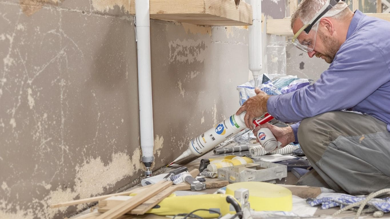 Person waterproofing basement wall with tools and materials.